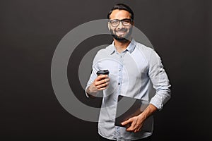 A relaxed Indian businessman with a broad smile holds a takeaway coffee and a laptop