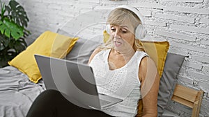 Relaxed middle age blonde woman comfortably sitting on a bedroom bed, engaging in a serious online video call conversation