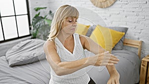 Relaxed middle age blonde woman, sitting comfortably on bed, applying skin treatment cream on arm, while unwinding in serene