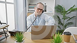 Relaxed middle age businessman engaging in a serious phone conversation while working on his laptop at the office