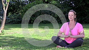 Relaxed middle aged woman in her forties outside cross legged practicing yoga in prayer