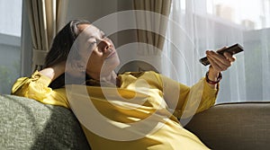 Relaxed middle aged woman watching tv at home, sitting on couch with remote control