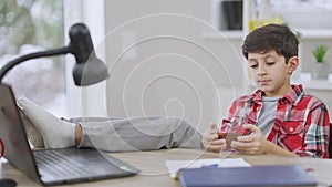 Relaxed Middle Eastern little boy sitting with feet on table and messaging online on smartphone. Bored schoolboy