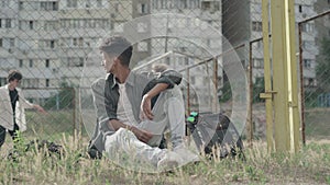 Relaxed mixed-race guy resting at mesh fence outdoors as Caucasian teenagers playing football at the background