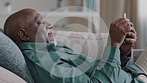 Relaxed old African American 60s bald bearded biracial man lie on soft comfortable couch at living room spend lazy