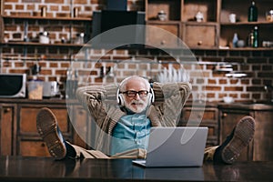 Relaxed old man in headphones using laptop with feet