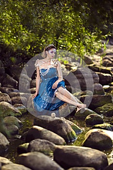 Relaxed Positive Lady Posing As Sea Mermaid  in Artistic Blue Dress And Strasses on Face Sitting in Wet Dress on Rocky Shore