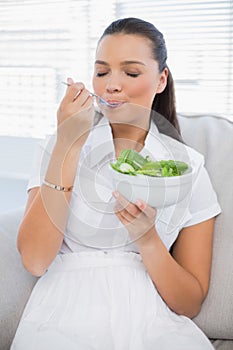 Relaxed pretty woman eating healthy salad sitting on sofa