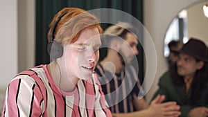 Relaxed redhead man listening to music in headphones with blurred friends talking at background. Portrait of carefree