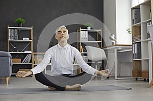 Relaxed senior businessman meditates sitting in lotus position on sports mat in office.