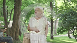 Relaxed senior Caucasian woman strolling in sunny park on summer day. Portrait of carefree female retiree walking