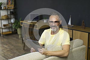 Relaxed senior man using digital tablet and smiling while sitting on the sofa at home