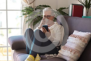 A relaxed senior woman sitting on the sofa at home drinking a coffee cup and looking at her smart phone - attractive woman with