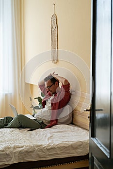 Relaxed serious Caucasian man attentively looks at laptop screen sits on bed with hands behind head