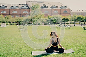 Relaxed slim brunette Caucasian woman sits in lotus pose on fitness mat has perfect body shape does yoga and meditation wears