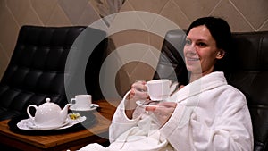 Relaxed smiling woman drinking tea resting lying in armchair in spa sauna.