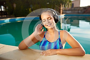 Relaxed smiling woman listening to music in headphones bathing in swimming pool. Blonde girl enjoys favourite song with