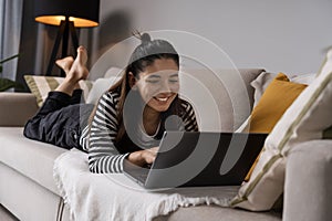 Relaxed study. Attractive student lying on sofa bed cheerfully working on laptop