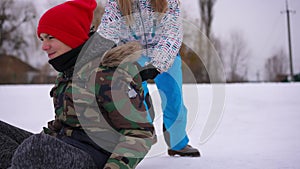 Relaxed teen boy sitting on green sled as girl pushing friend leaving. Portrait of joyful Caucasian friends having fun