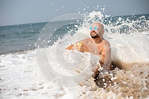 Relaxed vacationist in sunglasses resting on shore covered by sea wave, holding glass of drink in hand and smiling.