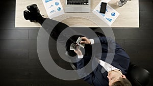 Relaxed wealthy businessman counting money, sitting with feet on table, top view