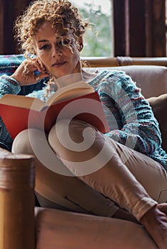 Relaxed woman alone at home sitting on a chair and reading a red book - female people indoor leisure relax activity read and enjoy