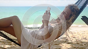 Relaxed woman browsing smartphone while lounging in hammock on beach