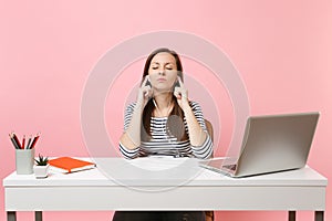 Relaxed woman with closed eyes in earphones listen music sitting and work at white desk with contemporary pc laptop