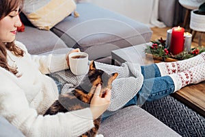 Relaxed woman with cup of hot drink petting cat on the sofa at home while watching movie, TV with christmas decoration