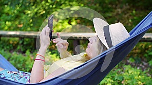 Relaxed woman lying in garden hammock using mobile phone in summer countryside. Happy woman in hat resting in summer