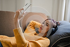 Relaxed woman lying on sofa, listening to music with headphones and using smartphone