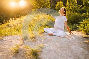 Relaxed woman sitting in lotus pose and doing bend in back outside in park evening on background of sunlight.