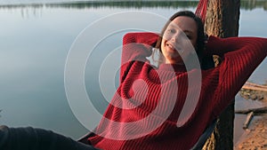 Relaxed woman smiling looking in camera lying in hammock near lake on nature.