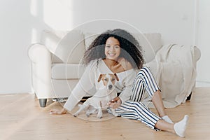 Relaxed young African American woman sits on floor in empty room near sofa with pedigree dog, enjoys sweet moment at home, move in