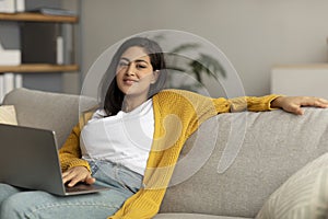 Relaxed young arab lady working from home, sitting on couch and using laptop, enjoying her weekend, surfing on internet