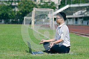 Relaxed young Asian business man with laptop doing yoga position on the green grass of stadium