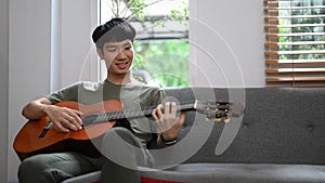 Relaxed young asian man playing acoustic guitar, spending leisure time in bright living room