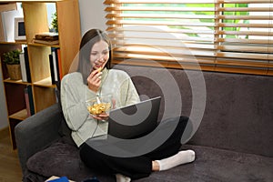 Relaxed young caucasian woman eating popcorn and watching movie on a laptop at home