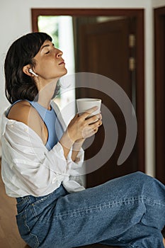 Relaxed young caucasian woman holding cup, listening to music with her eyes closed sitting indoors.