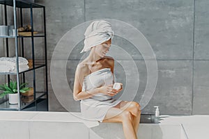 Relaxed young European woman applies moisturizer cream on legs after taking bath sits wrapped in towel in bathroom, enjoys beauty