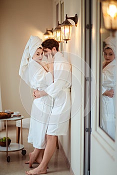 Relaxed young female and male wears white bathrobe, embrace each other, feel relief after taking bath, stand in hotel