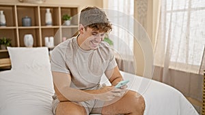 Relaxed young hispanic man sitting in cozy bedroom, all smiles as he celebrates his online win from the comfort of his bed, using