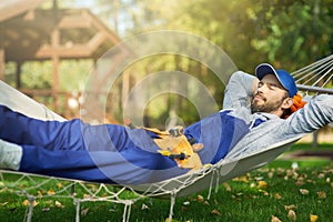 Relaxed young male builder wearing blue overalls and cap taking a break, lying in a hammock outdoors with eyes closed on