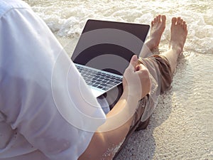 Relaxed young man with laptop sitting on the sandy beach with soft waves. Internet of things concept