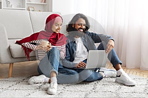 Relaxed young muslim couple sitting on floor with laptop and coffee mug