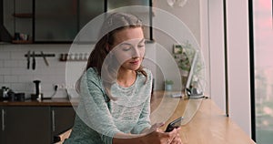 Relaxed young smiling woman using smartphone in modern kitchen.