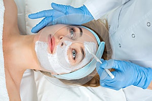 Relaxed young woman is getting facial skin care treatment at beauty salon. Beautician is touching brush with clay to face girl