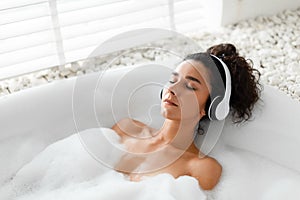 Relaxed young woman listening to calming music in wireless headphones, lying in bubble bath at home, copy space