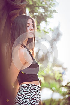 Relaxed young woman outdoors in nature with closed eyes. She leaning against the trunk of a tree. Warm evening lights