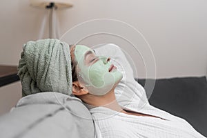 Relaxed young woman with peeling clay mask on face lying on comfortable sofa spa procedures